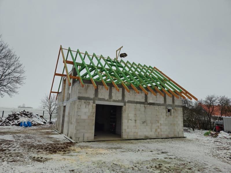 Surowy budynek z betonu komórkowego w trakcie budowy, z zieloną konstrukcją więźby dachowej, na zasnieżonym terenie.