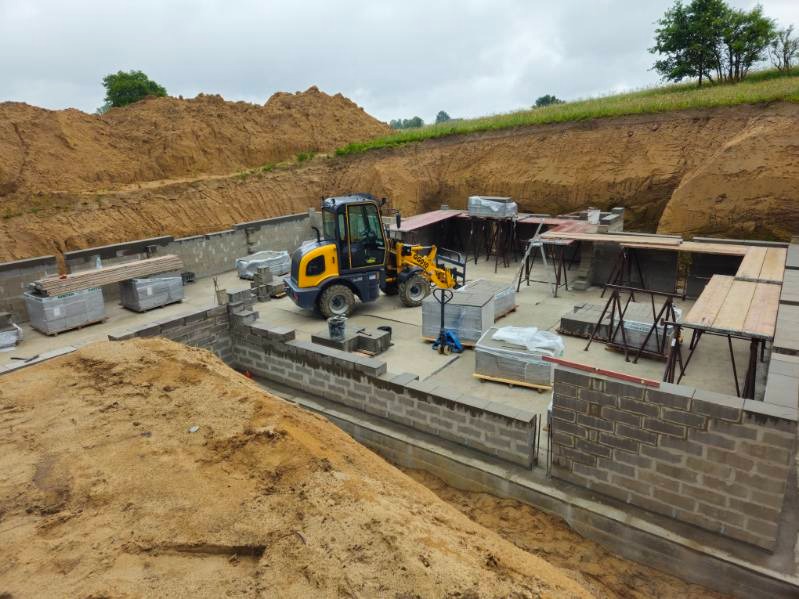 Budowa fundamentów domu z bloczków betonowych, widoczny żółty miniładowarka, wykop pod fundamenty i przygotowane podpory pod szalunek.