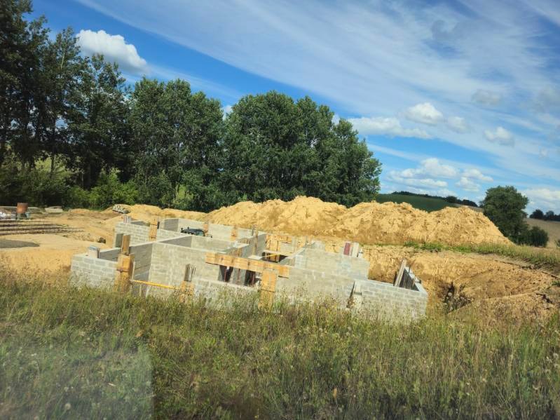 Wznoszone z bloczków fundamenty domu jednorodzinnego na tle hałdy piasku i letniego krajobrazu z błękitnym niebem i chmurami.