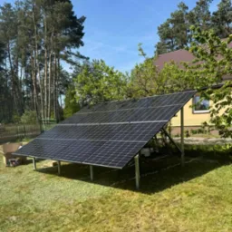 Instalacja fotowoltaiczna na gruncie z ośmioma panelami Jinko Solar 480W i inwerterem Sofar Solar 5kW, widoczne narzędzia i karton po panelach, tło stanowią drzewa i budynek z brązowym dachem.