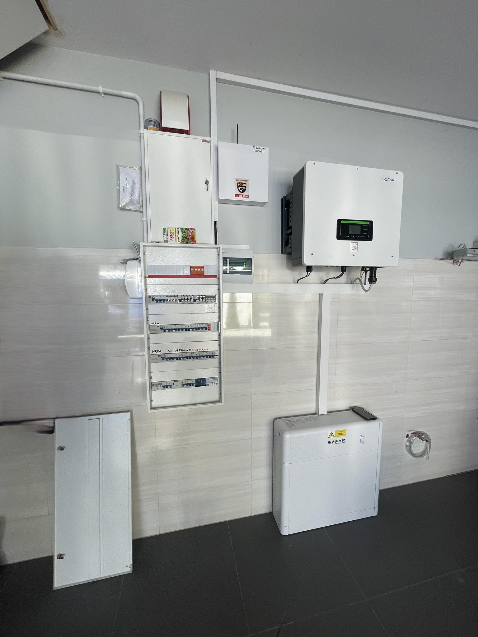 Zamontowany inwerter hybrydowy Sofar Solar połączony z magazynem energii Sofar Solar, widoczna rozdzielnica elektryczna i okablowanie na ścianie z jasnych płytek ceramicznych.