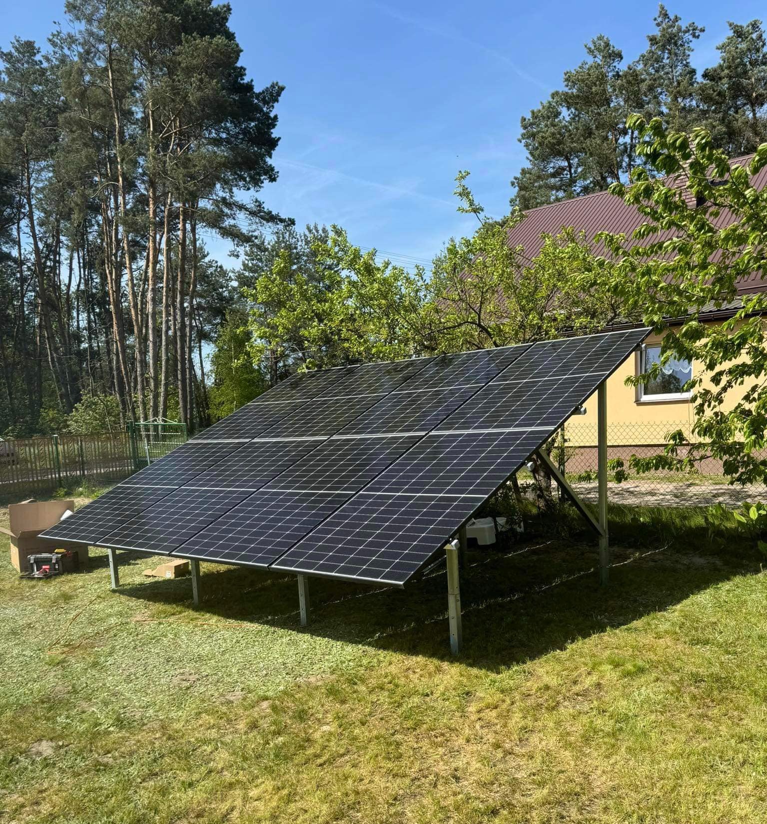 Instalacja fotowoltaiczna na gruncie z ośmioma panelami Jinko Solar 480W i inwerterem Sofar Solar 5kW, widoczne narzędzia i karton po panelach, tło stanowią drzewa i budynek z brązowym dachem.