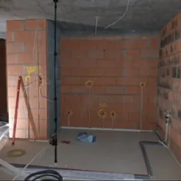 Surowy stan wnętrza z ceglanymi ścianami, widoczne punkty instalacji elektrycznej oznaczone żółtym markerem, wystające przewody, poziomica oparta o ścianę, oraz narzędzia i materiały budowlane...