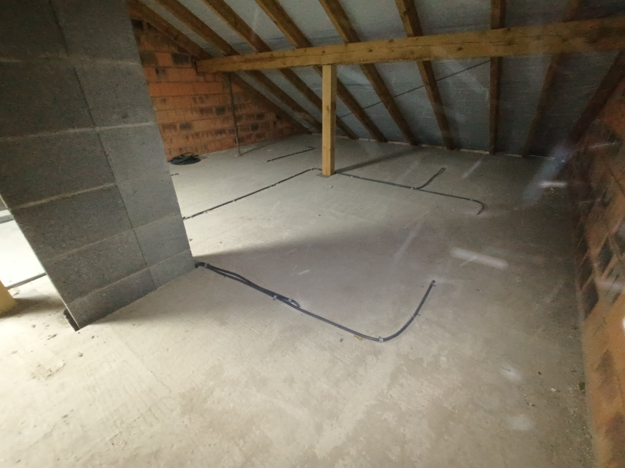 Surowy strych w trakcie adaptacji: widoczne kable elektryczne ułożone na betonowej podłodze, ceglana ściana i drewniana konstrukcja dachu.