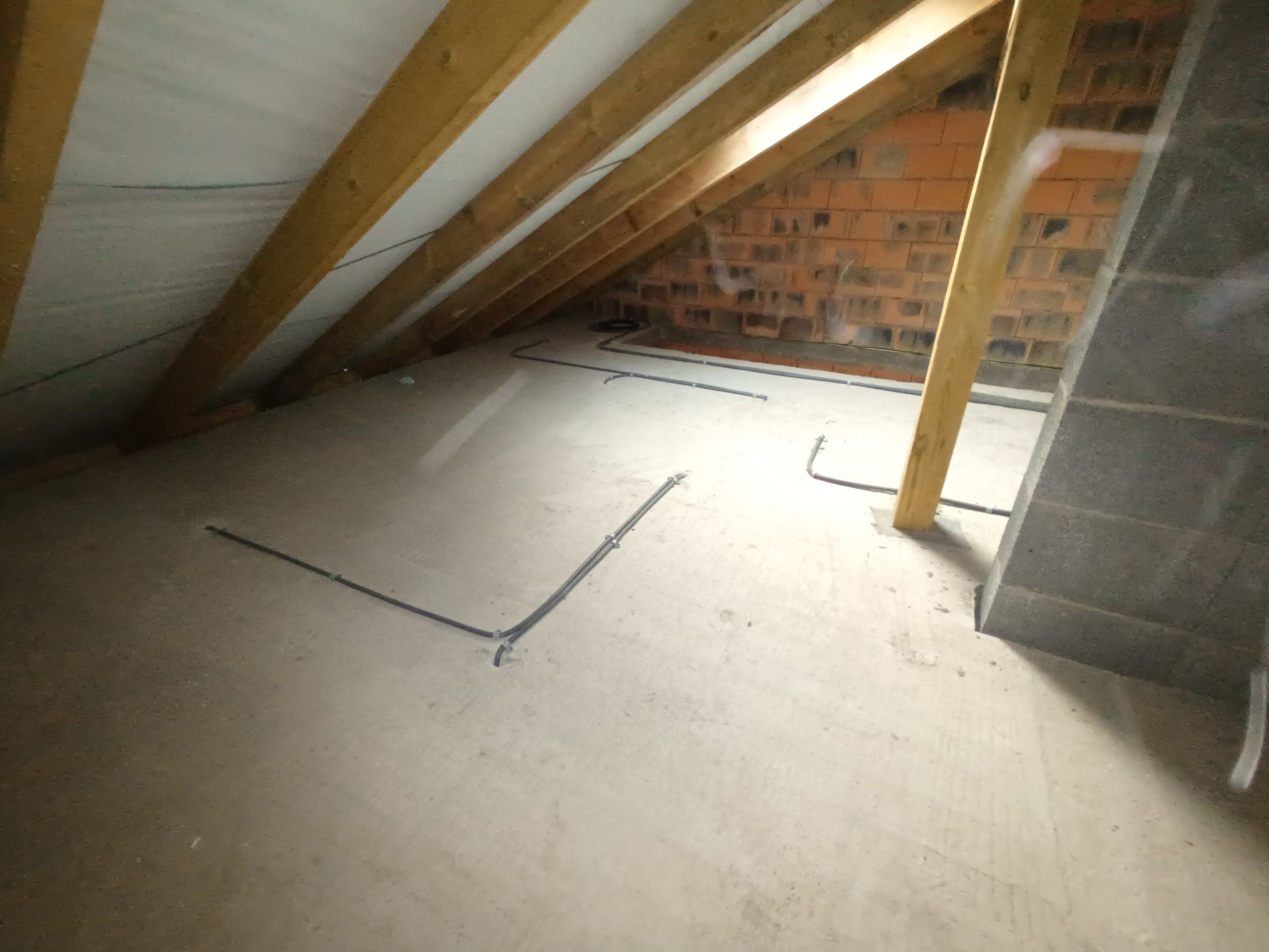 Instalacja przewodów elektrycznych w przestrzeni poddasza z widoczną konstrukcją drewnianą dachu i murem z cegły.