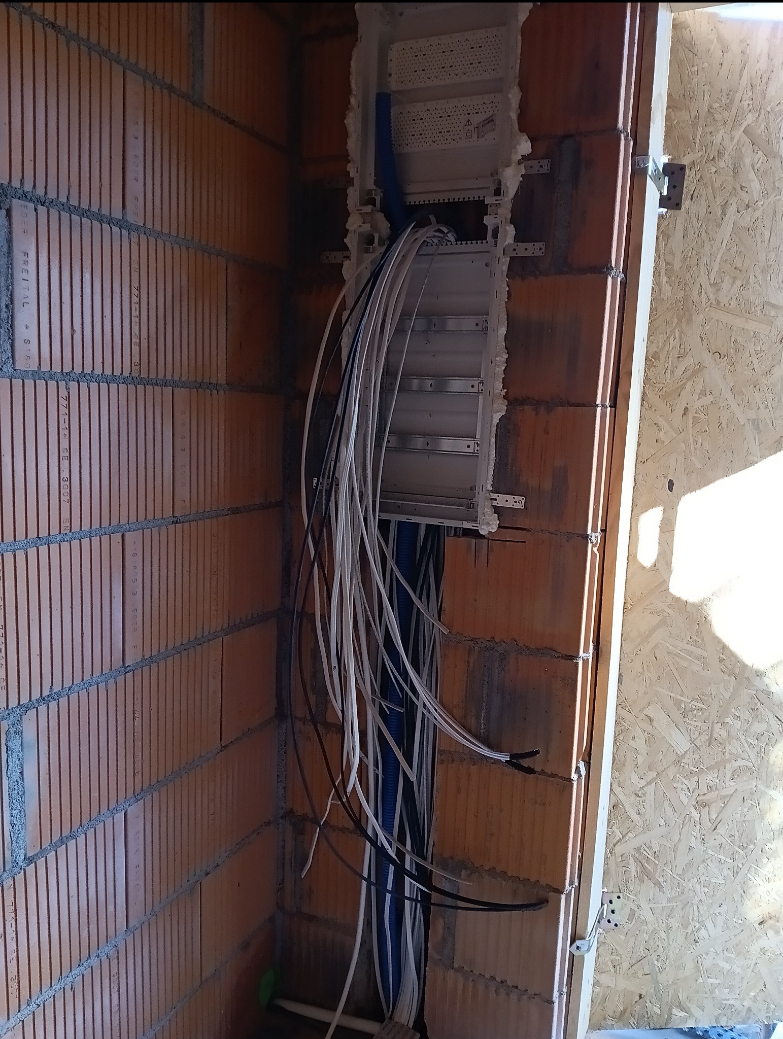 Rozdzielnica elektryczna zamontowana w ścianie z cegły dziurawki w trakcie instalacji, widoczne liczne przewody w kolorach białym, czarnym i niebieskim, wystające ze skrzynki i prowadzone w dół...