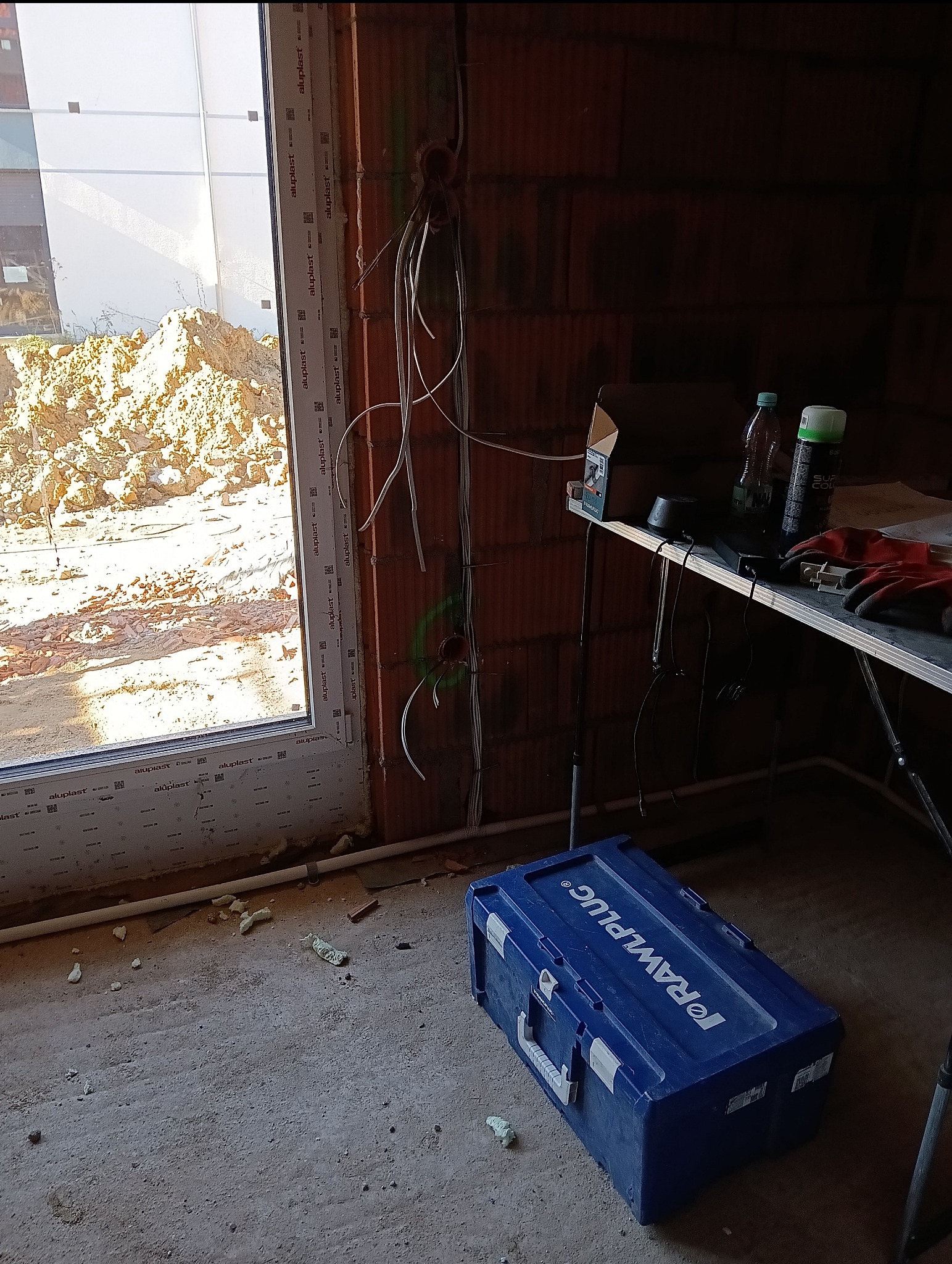 Wnętrze w trakcie prac wykończeniowych; widoczne wystające przewody elektryczne ze ściany z cegły, okno z widokiem na ziemię oraz skrzynka narzędziowa Rawlplug na podłodze.