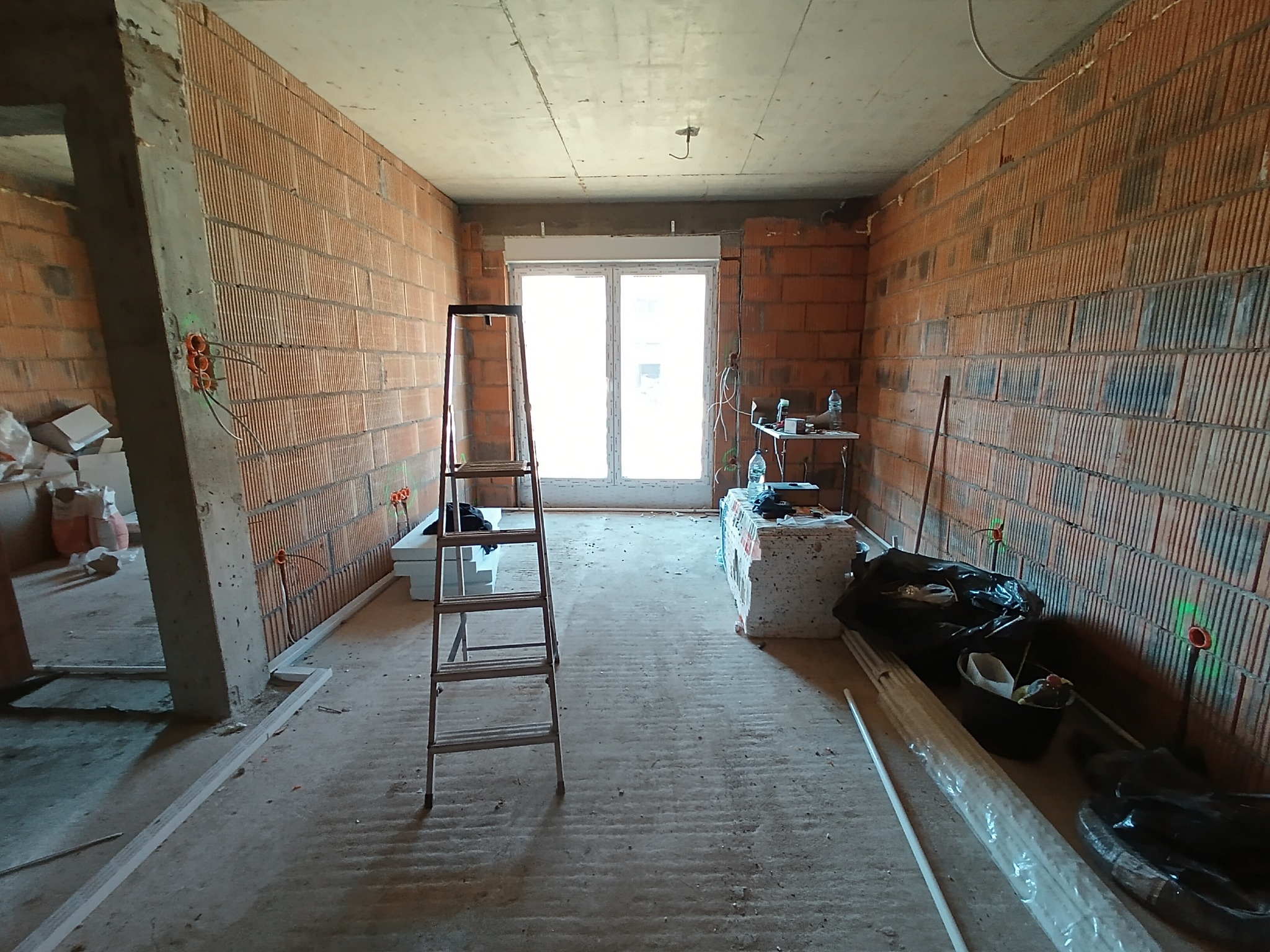 Surowy stan wnętrza z ceglanymi ścianami, widoczne puszki elektryczne, drabina aluminiowa i materiały budowlane przygotowane do dalszych prac wykończeniowych.