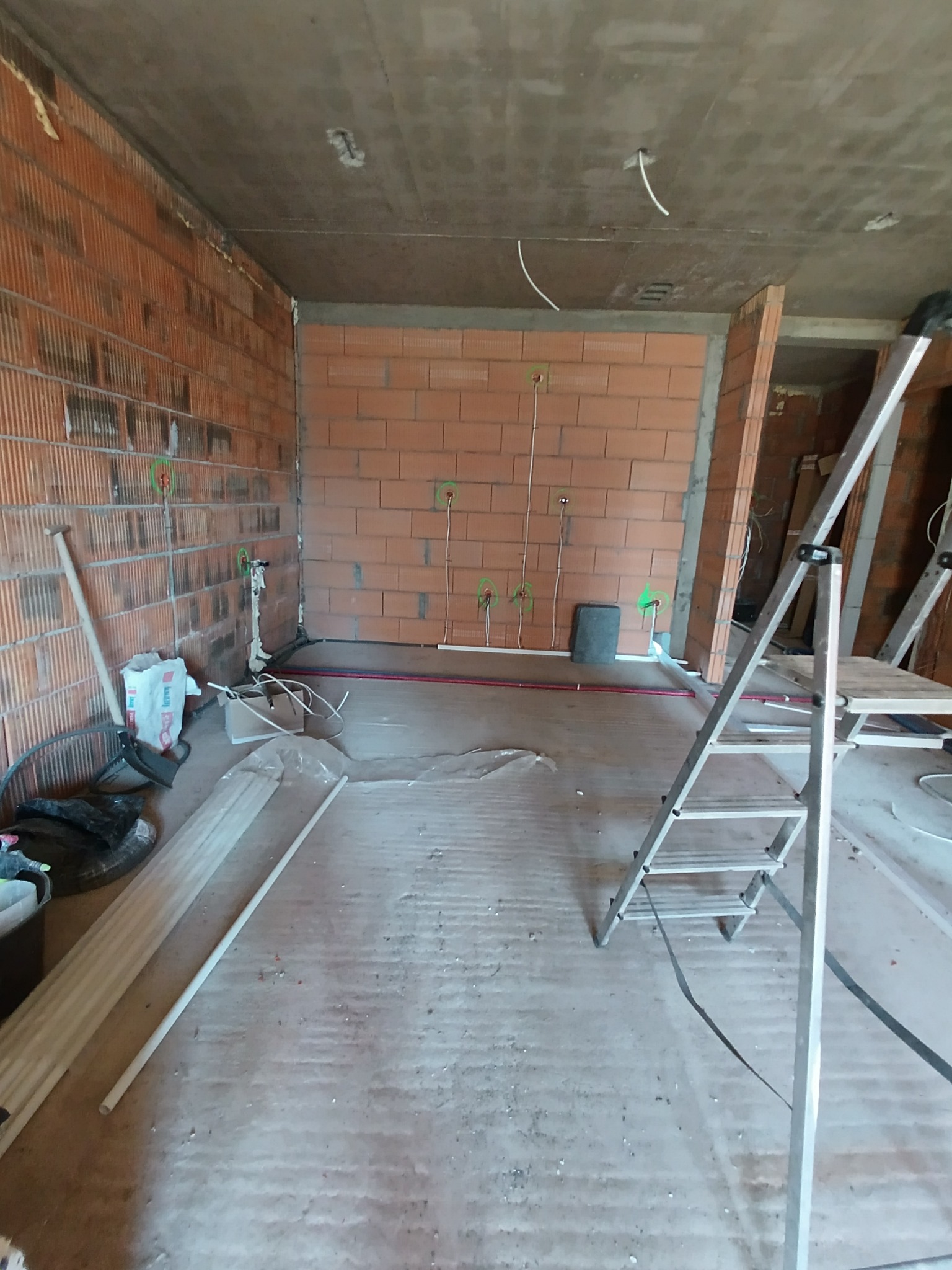 Wnętrze w trakcie remontu w Bolesławcu: surowe ściany z cegły, instalacje elektryczne wyprowadzone ze ścian, drabina aluminiowa, worki z zaprawą i narzędzia budowlane.
