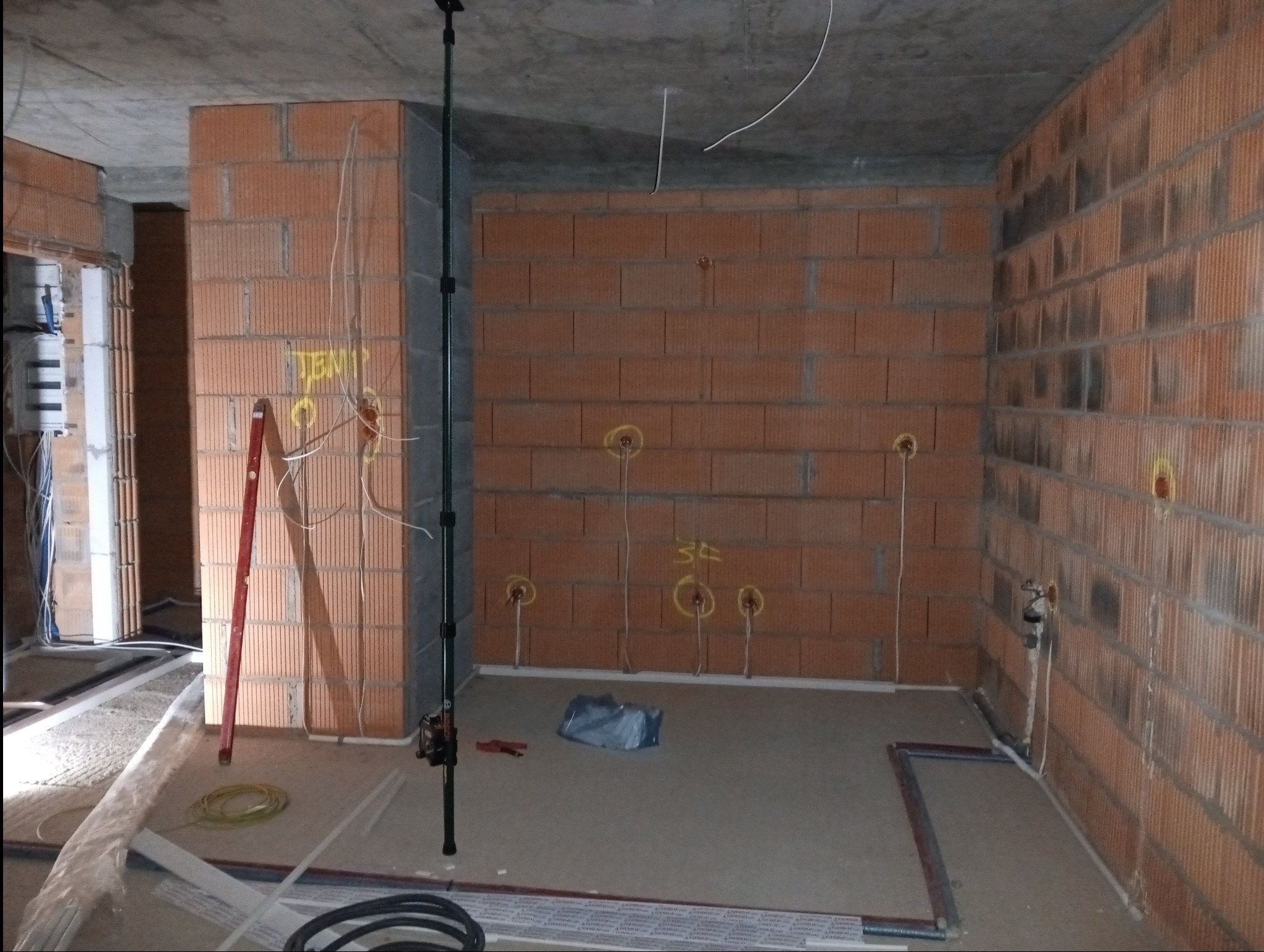 Surowy stan wnętrza z ceglanymi ścianami, widoczne punkty instalacji elektrycznej oznaczone żółtym markerem, wystające przewody, poziomica oparta o ścianę, oraz narzędzia i materiały budowlane...