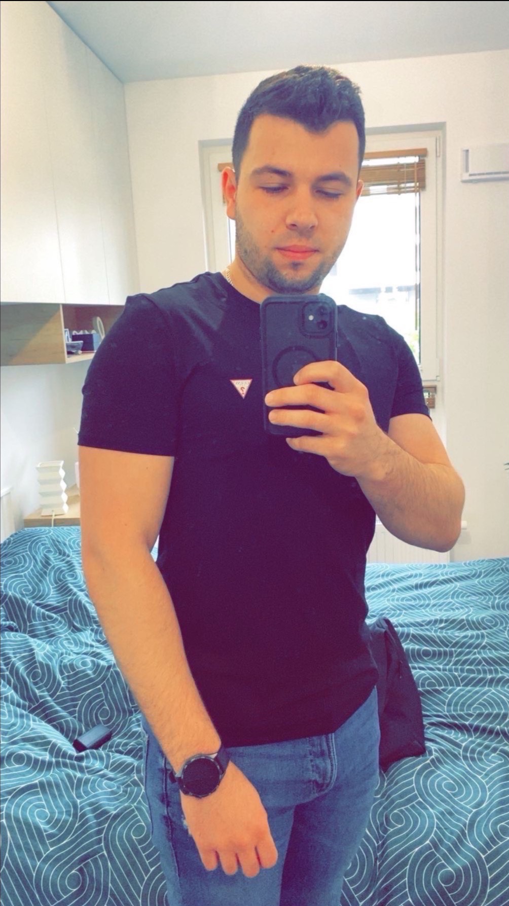 Mężczyzna robi selfie w pokoju, w tle widoczne białe ściany, okno z żaluzjami i fragment łóżka z niebieską pościelą w białe wzory.