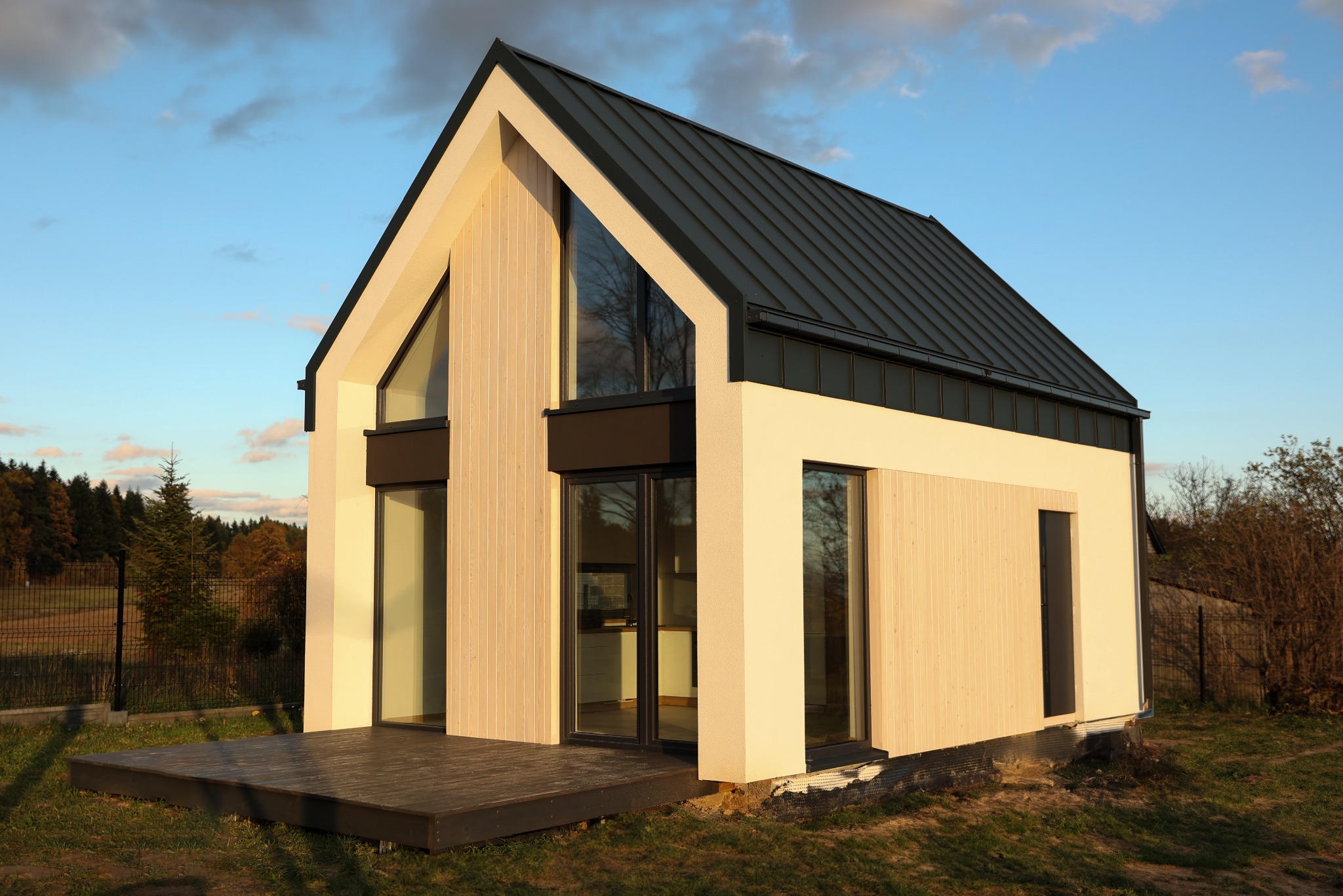 Nowoczesny, parterowy dom z dachem dwuspadowym i dużymi przeszkleniami, wykończony jasnym drewnem na tle jesiennego krajobrazu.