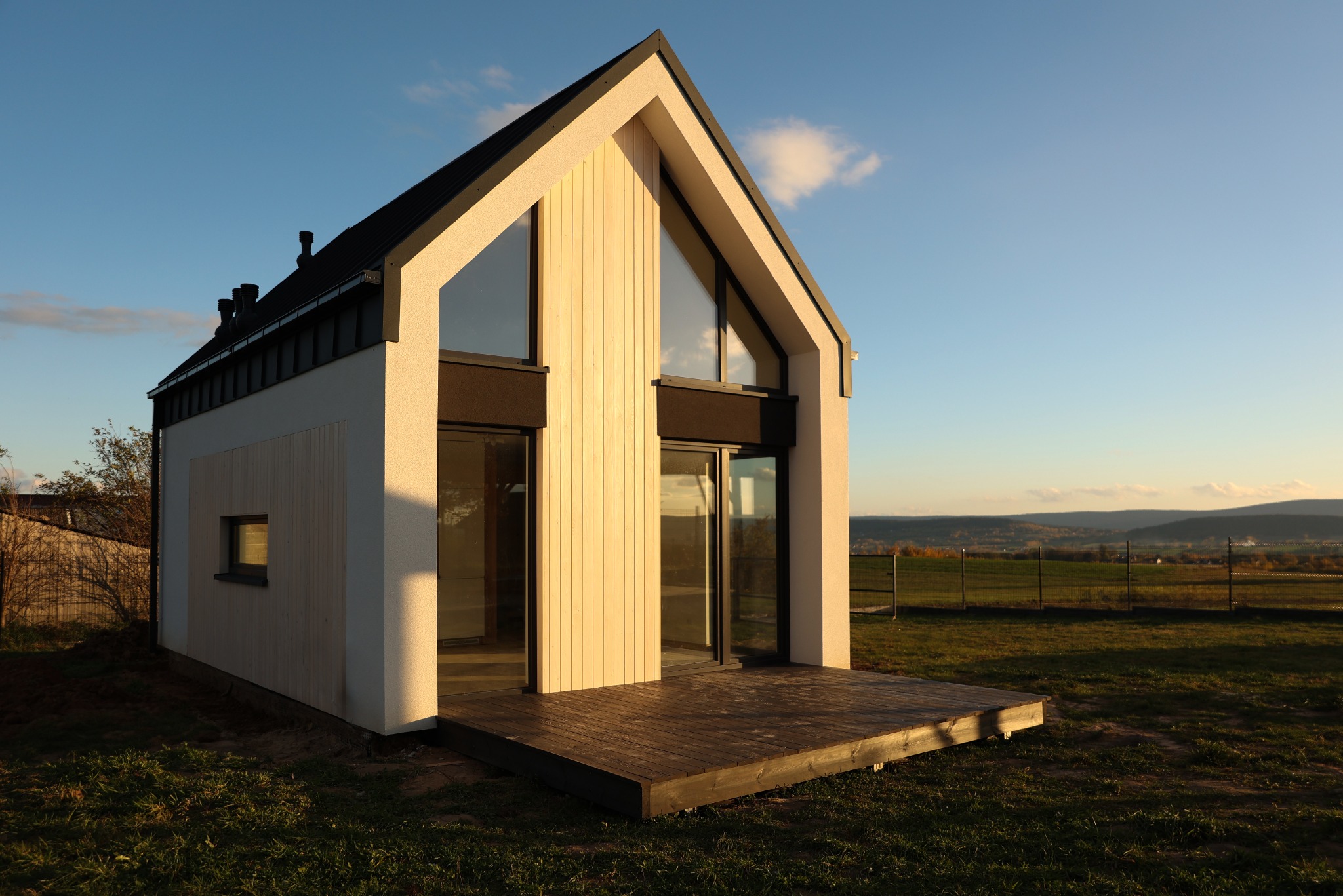 Nowoczesny dom z białymi ścianami i ciemnym dachem, z dużymi oknami i drewnianym tarasem, na tle zielonej trawy i pagórkowatego krajobrazu o zachodzie słońca.