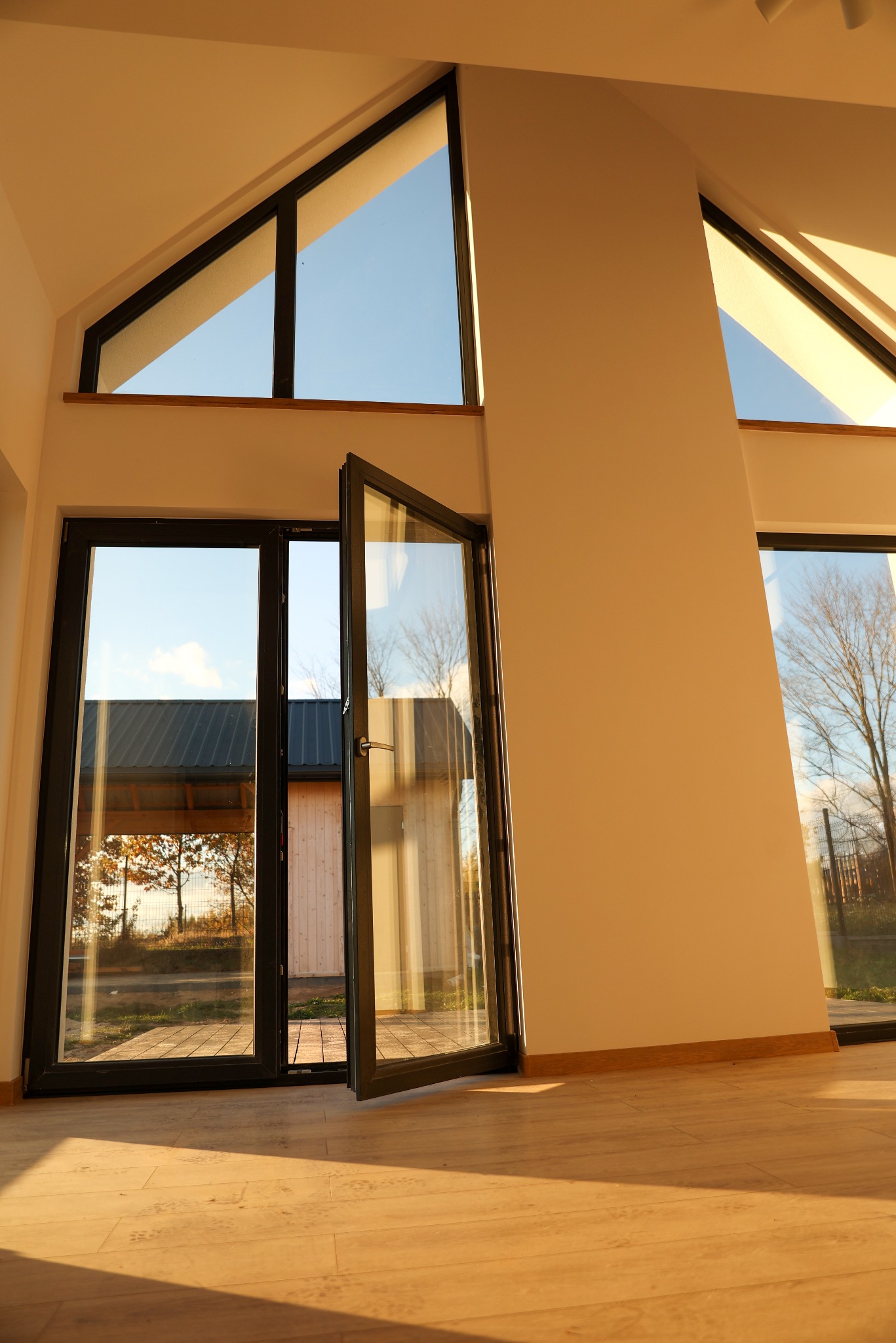 Wnętrze domu w stanie deweloperskim z dużymi oknami trójkątnymi i otwartymi drzwiami balkonowymi z czarnymi ramami, widok na ogród i sąsiedni budynek.