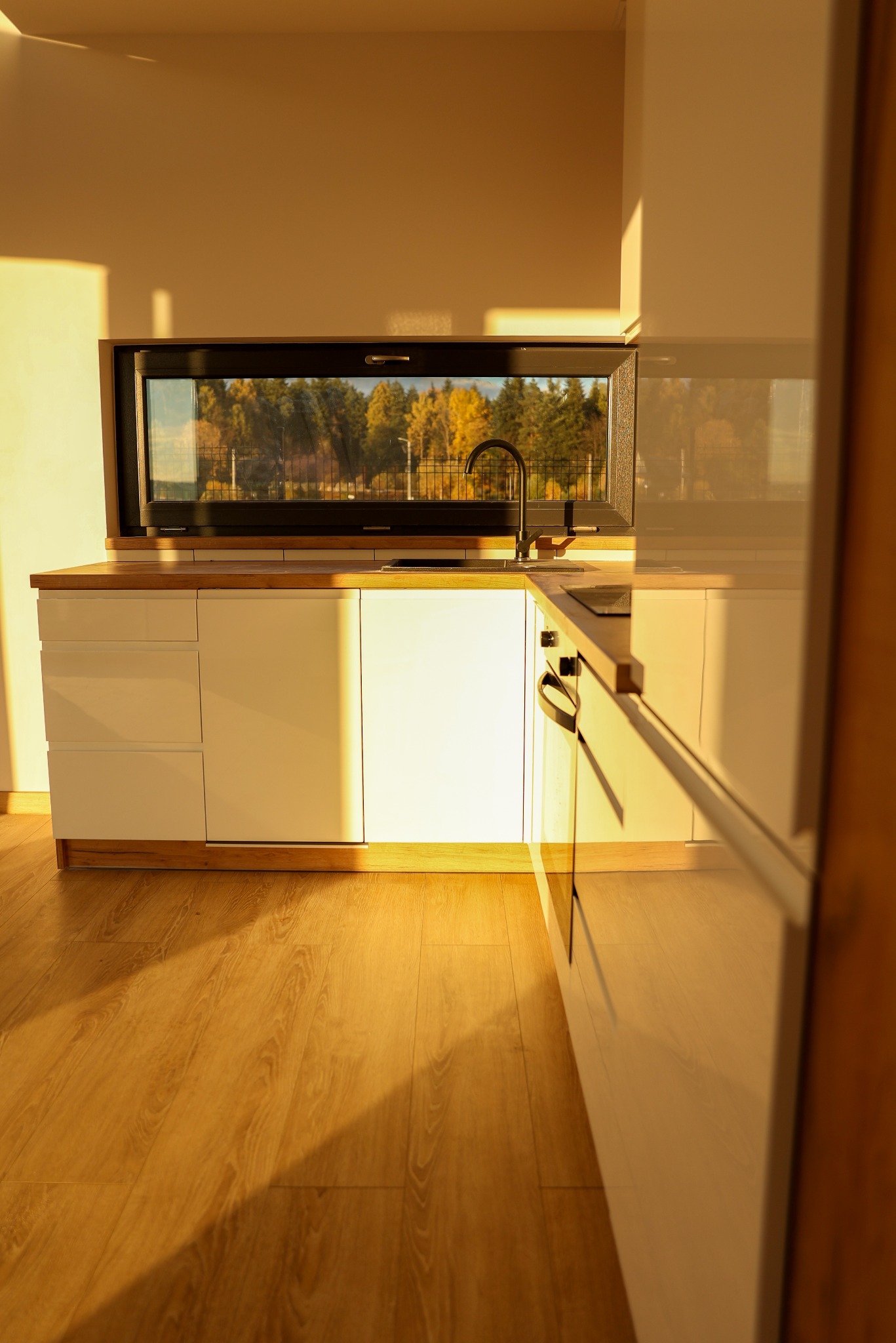 Nowoczesna kuchnia z białymi szafkami i drewnianym blatem, czarna bateria i okno z widokiem na jesienny las, oświetlona ciepłym, popołudniowym światłem.