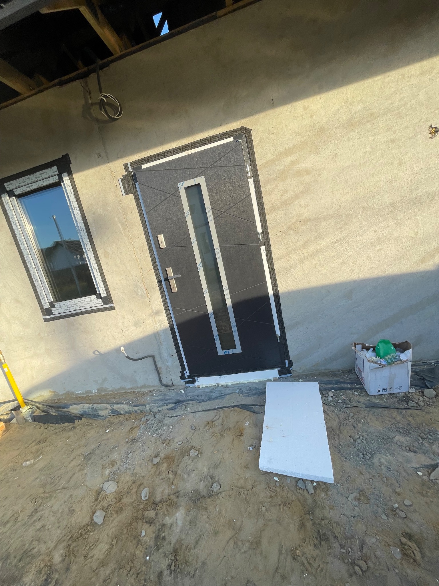 Nowo zamontowane, grafitowe drzwi zewnętrzne z długim, pionowym przeszkleniem, widoczne obok okna w szarej ramie, na tle surowej elewacji i gruntu przed budynkiem.