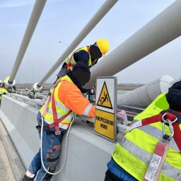 FW-EXPERT - Pracownicy w odblaskowych kamizelkach i kaskach, zabezpieczeni linami, montują instalację na wysokości, obok znaku ostrzegawczego 'Niebezpieczeństwo Upadku'.