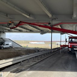 FW-EXPERT - Pracownik w koszu podnośnika teleskopowego, montujący instalację pod wiaduktem drogowym; w tle zaparkowany bus.