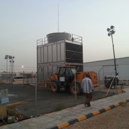 FW-EXPERT - Teren przemysłowy z dużą chłodnicą przemysłową, antenami i przenośnym masztem oświetleniowym, ogrodzony siatką i z przechodzącym mężczyzną.