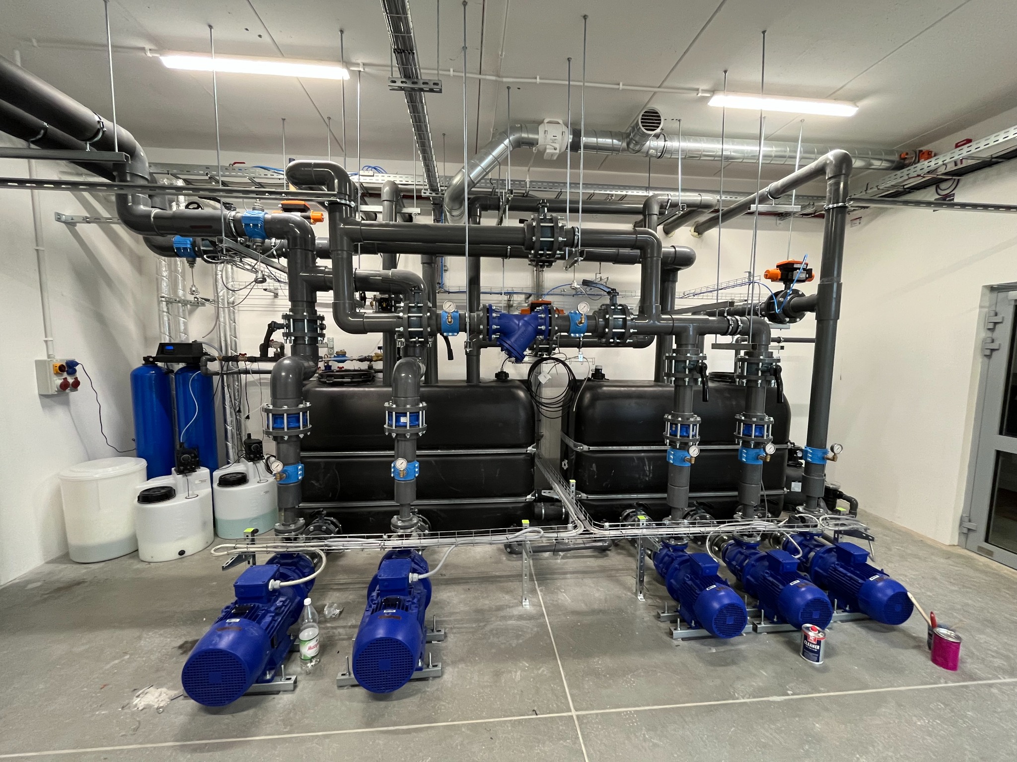 Kompleksowa instalacja chłodzenia wtryskarek z rurami, zbiornikami i pompami w przemysłowym pomieszczeniu, widoczne przewody i mocowania pod sufitem.