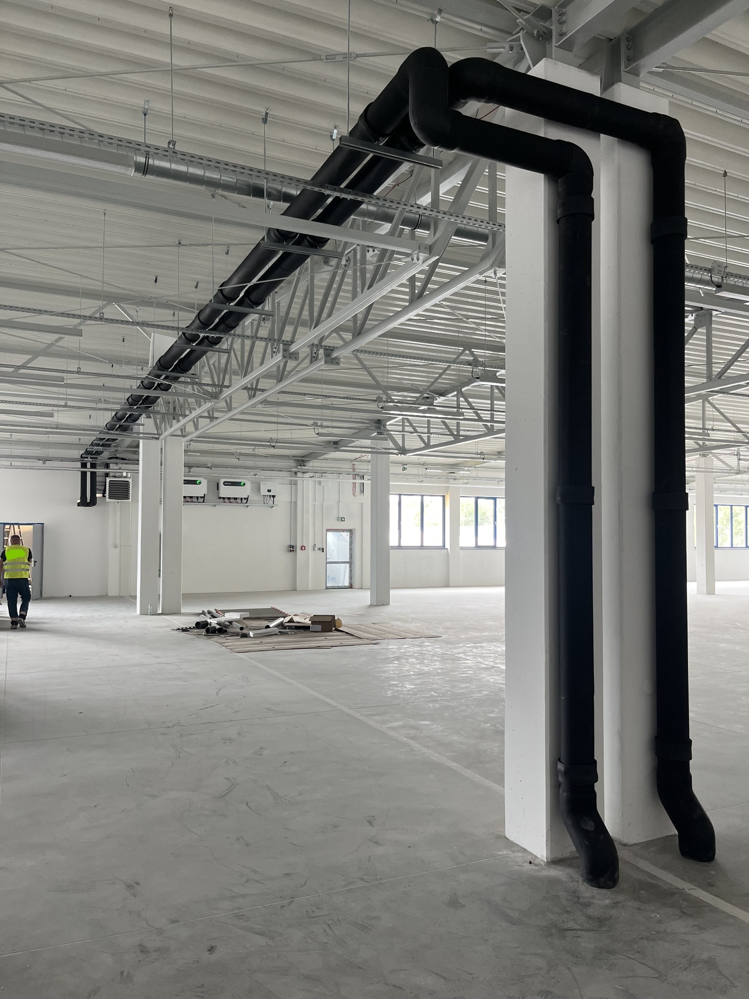 Czarny system rur chłodzących, biegnący wzdłuż sufitu i pionowo w dół po białym słupie w hali przemysłowej z widoczną metalową konstrukcją dachu i pracownikiem w odblaskowej kamizelce w tle.