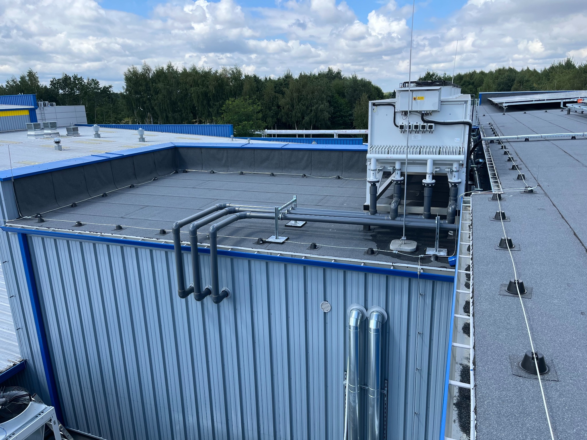 Instalacja chłodzenia przemysłowego na dachu budynku z blachy falistej, widoczne rury odprowadzające ciepło i agregat chłodniczy z elektryczną skrzynką sterującą.