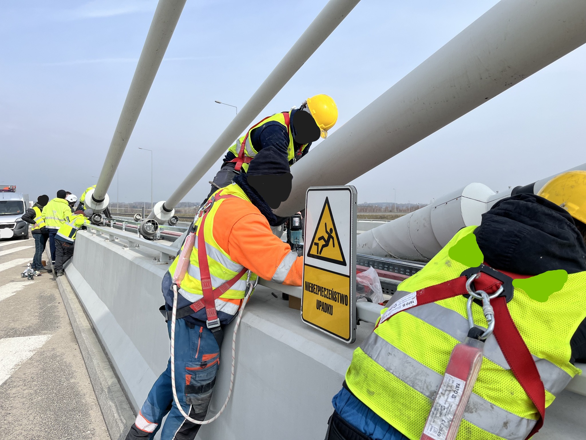 Pracownicy w odblaskowych kamizelkach i kaskach, zabezpieczeni linami, montują instalację na wysokości, obok znaku ostrzegawczego 'Niebezpieczeństwo Upadku'.