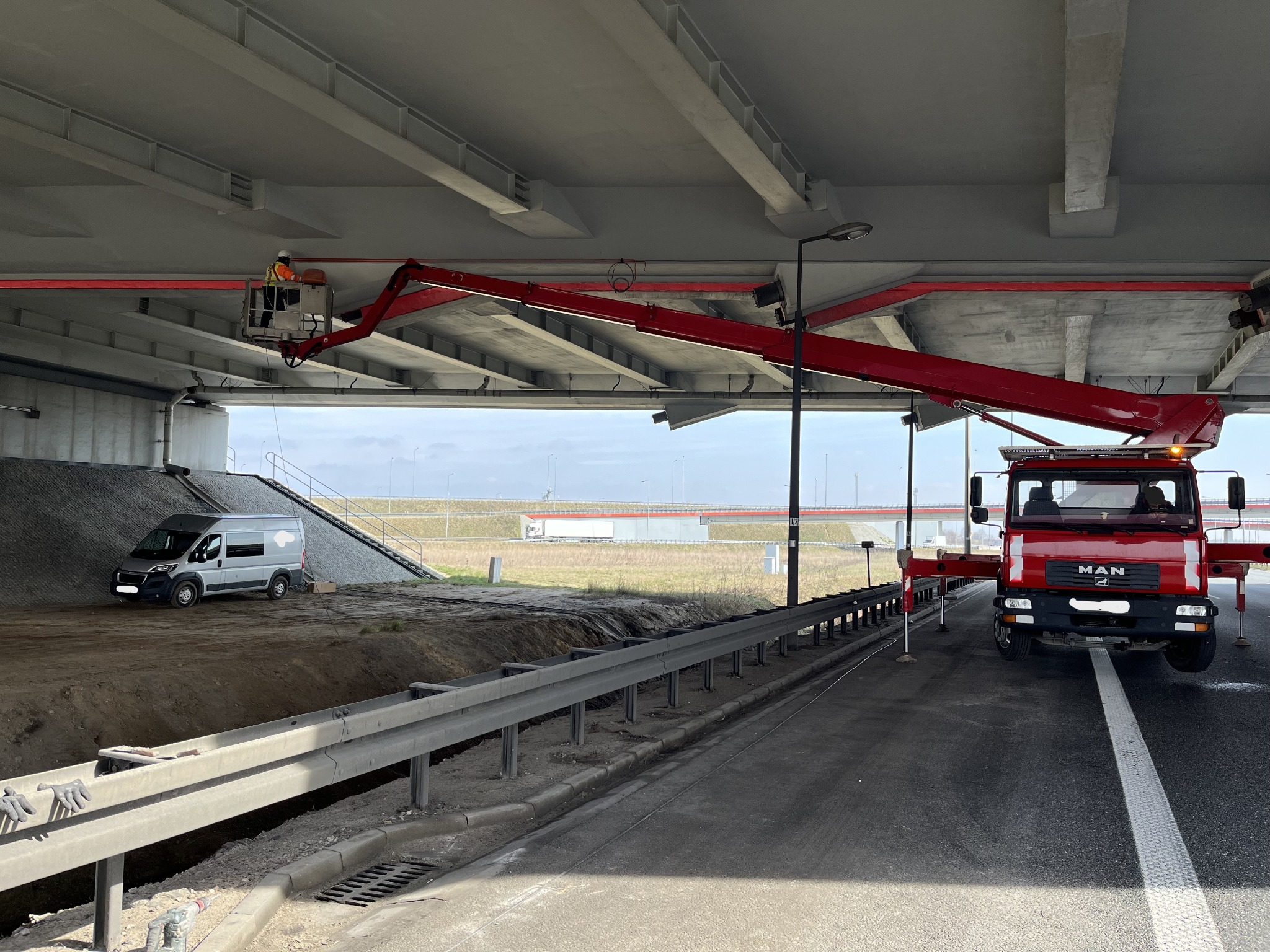 Pracownik w koszu podnośnika teleskopowego, montujący instalację pod wiaduktem drogowym; w tle zaparkowany bus.