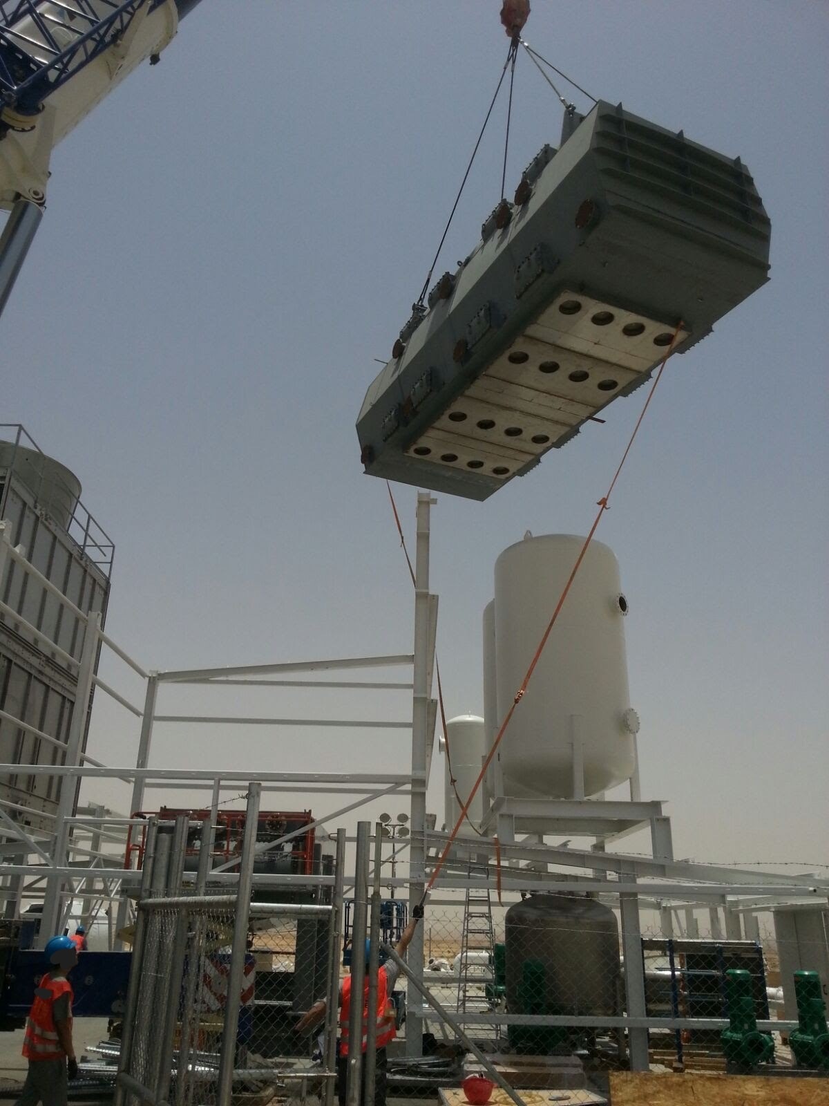 Kran podnosi duży, szary transformator z otworami wentylacyjnymi na placu budowy, widoczni pracownicy w kaskach i kamizelkach odblaskowych, zbiorniki ciśnieniowe w tle.