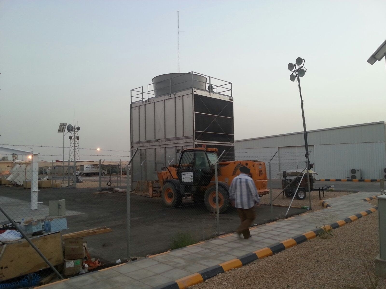 Teren przemysłowy z dużą chłodnicą przemysłową, antenami i przenośnym masztem oświetleniowym, ogrodzony siatką i z przechodzącym mężczyzną.