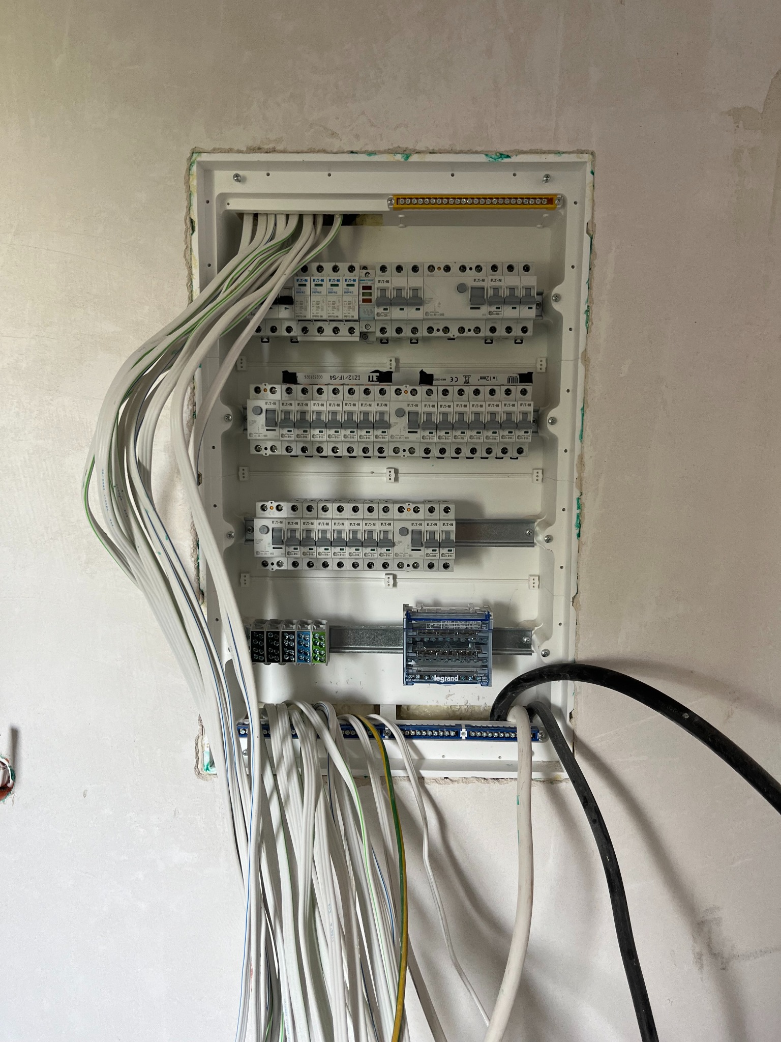 Rozdzielnica elektryczna w trakcie montażu, widoczne liczne przewody elektryczne, bezpieczniki ETI i moduły Legrand, ściana w stanie surowym.