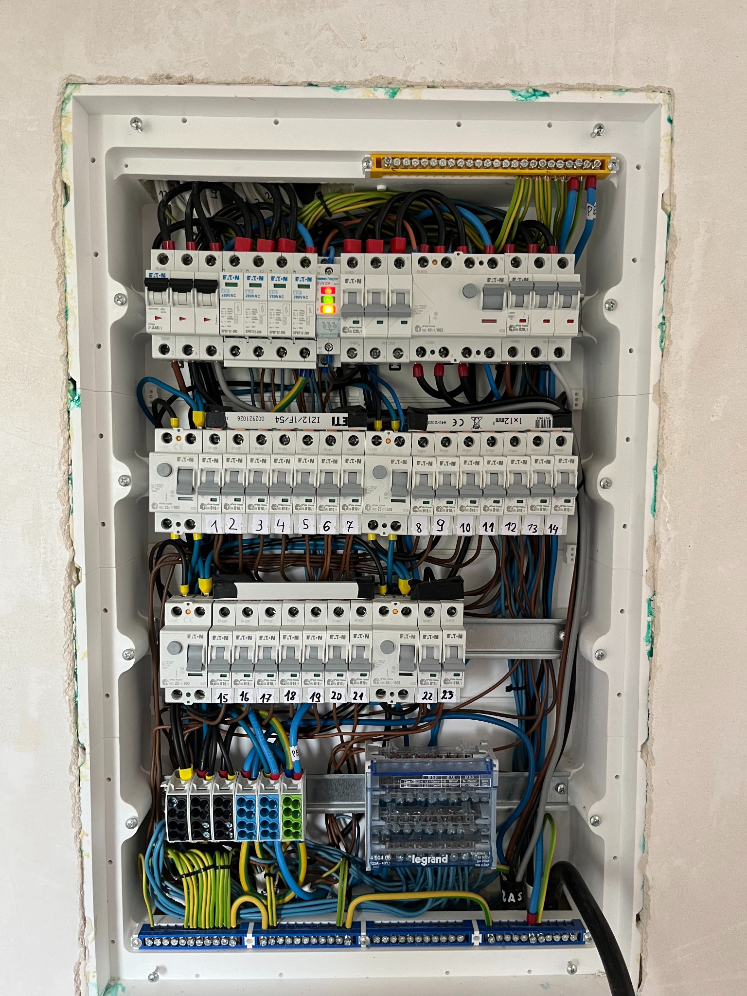 Otwarta skrzynka rozdzielcza z bezpiecznikami i okablowaniem, widoczne zabezpieczenia ETI i Eaton, oznaczone numery obwodów od 1 do 23, żółto-zielone przewody ochronne.