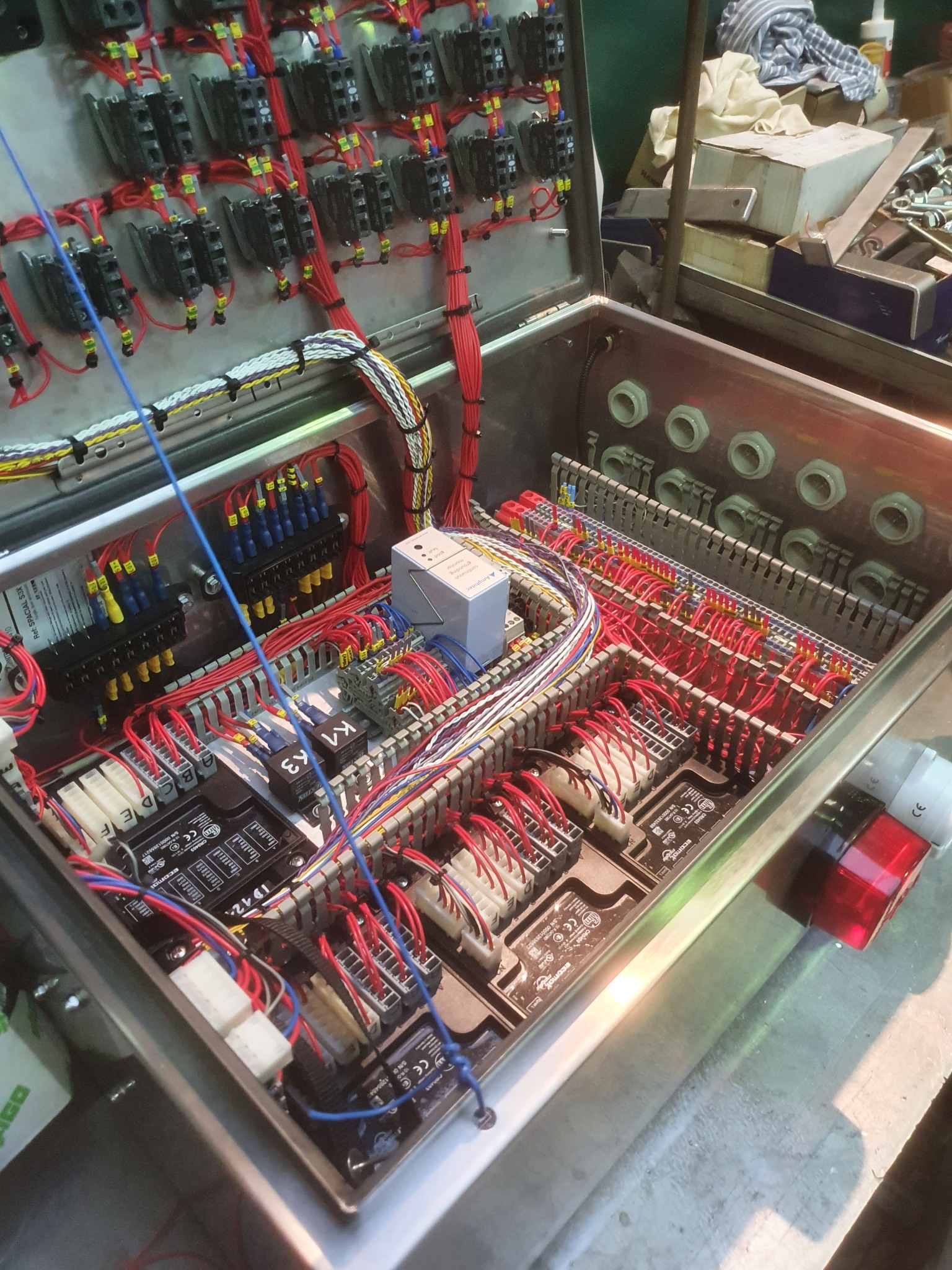 Otwarta metalowa szafa sterownicza z widocznymi licznymi przewodami, bezpiecznikami i innymi elementami elektrycznymi. W tle widoczne narzędzia i materiały.