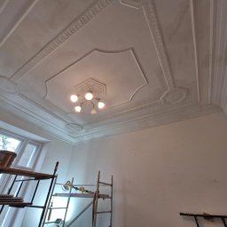 O&O Dominik Pawłowski - Wnętrze pokoju w trakcie odświeżania, widoczne rusztowania, wiadro z farbą i dekoracyjny sufit z oświetleniem.
