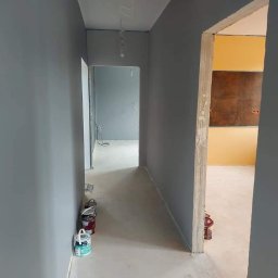 O&O Dominik Pawłowski - Korytarz w trakcie malowania na szaro, widoczne puszki z farbą przy ścianie, w tle otwarte drzwi do pomalowanego na żółto pokoju z brązowym panelem.