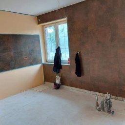 O&O Dominik Pawłowski - Pokój w trakcie prac wykończeniowych: ściany częściowo pomalowane na beżowo i brązowo z efektem rdzy, widoczne narzędzia malarskie i przewody elektryczne wystające ze ściany, okno z powieszonymi...