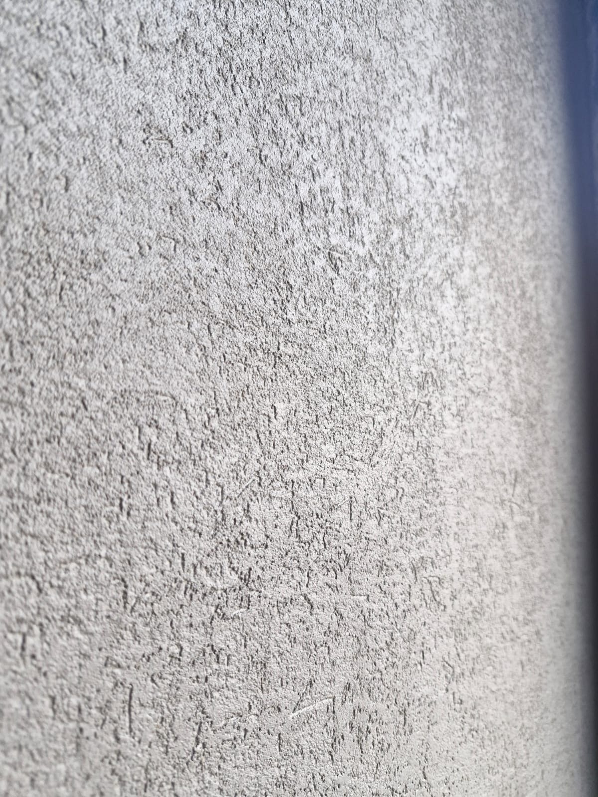 Szara ściana z wyraźną, chropowatą fakturą, oświetlona z boku, tworząca gradient cieniowania, ukazująca detale struktury.