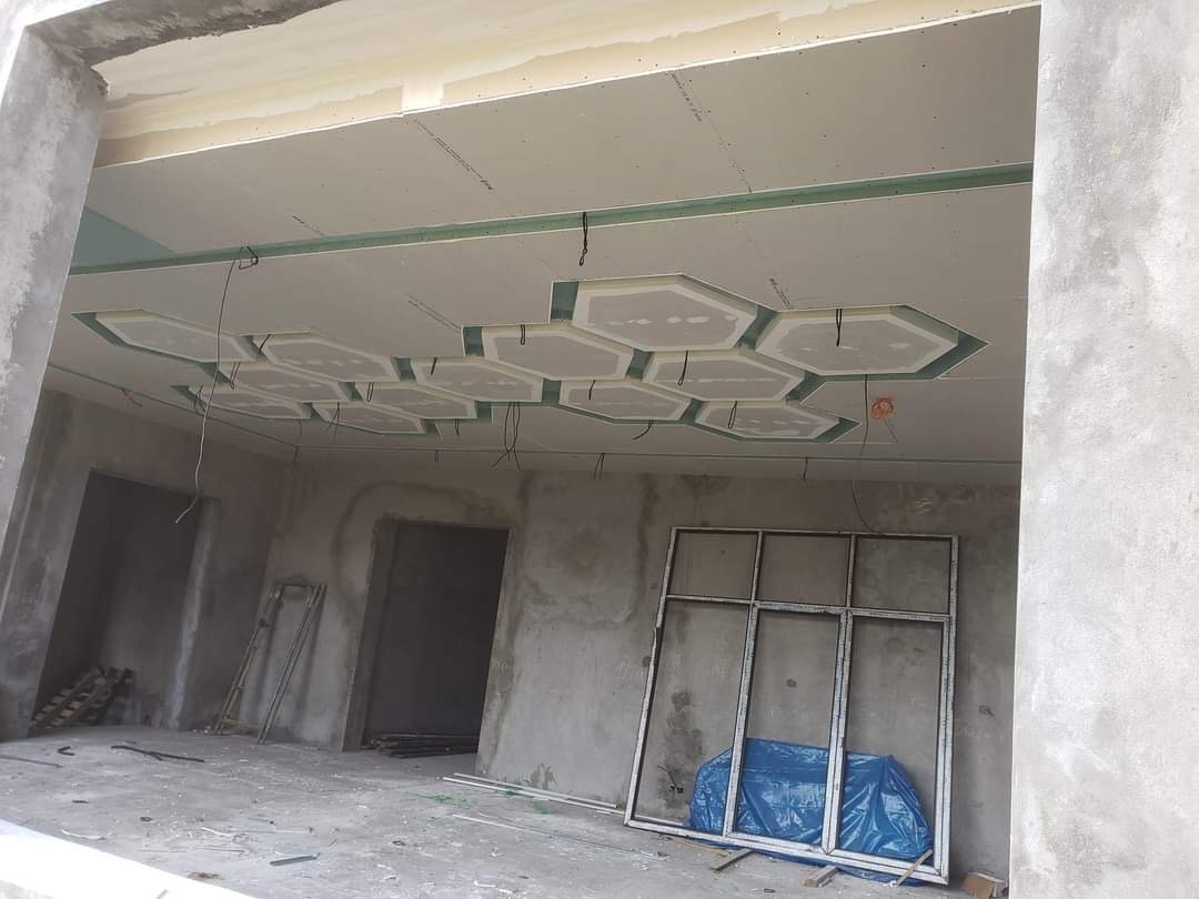 Wnętrze w trakcie wykończenia: sufit z płyt kartonowo-gipsowych z dekoracyjnymi heksagonami, widoczne kable, surowe ściany i okno zabezpieczone folią.