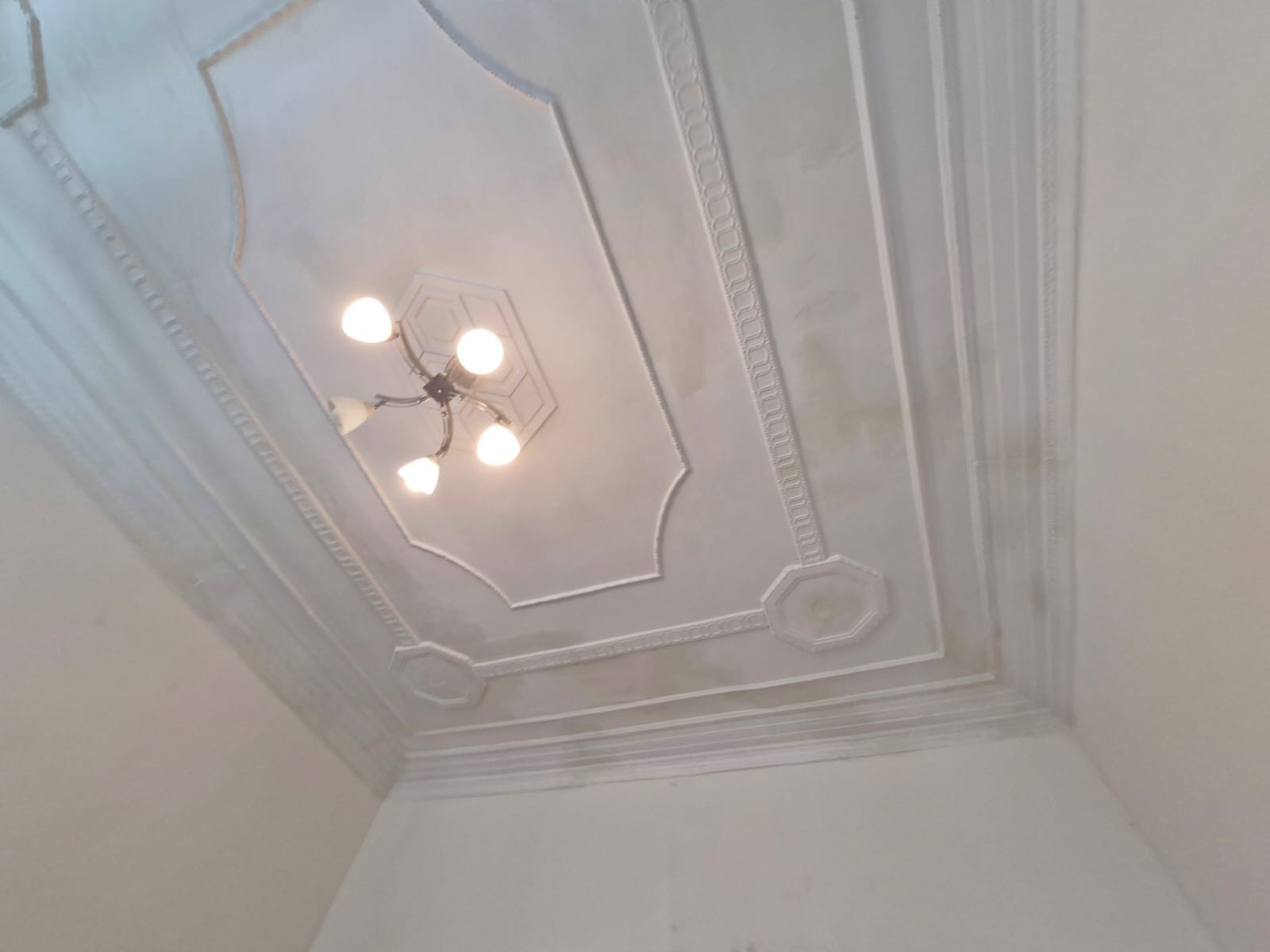 Biały sufit z dekoracyjnymi sztukateriami i centralnie umieszczonym żyrandolem z pięcioma żarówkami, widok z dołu pod kątem.