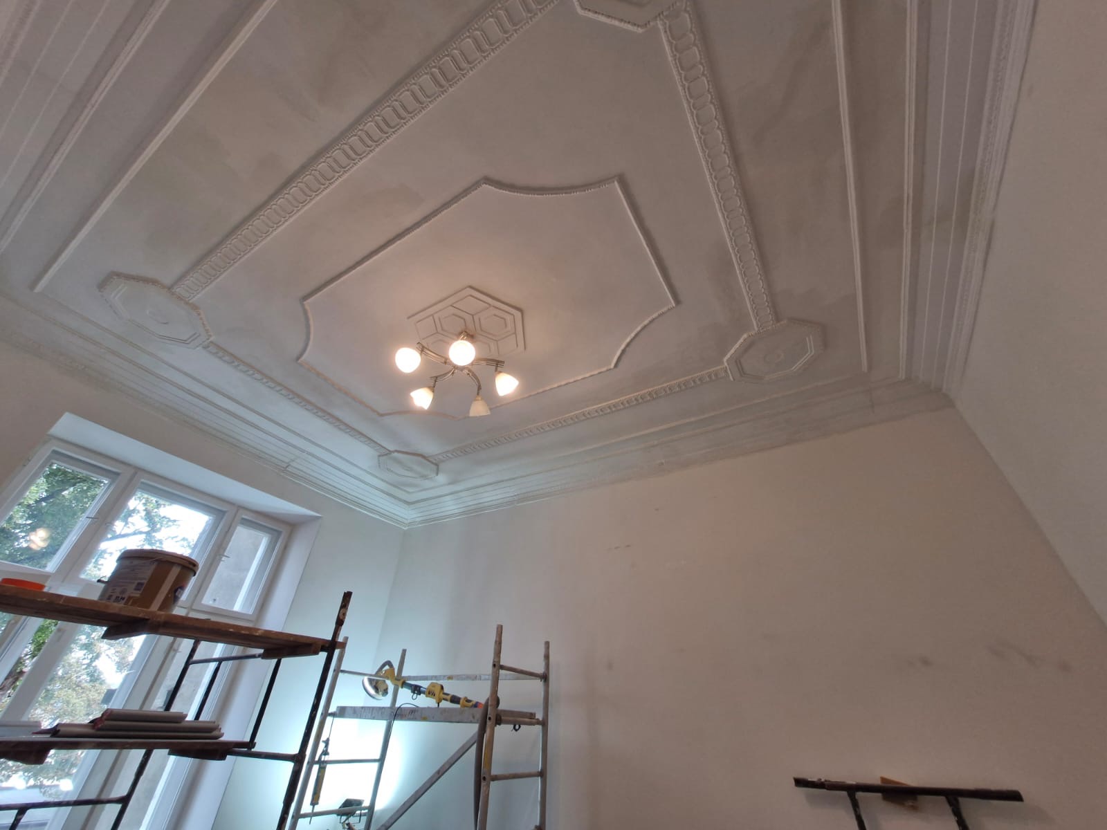 Wnętrze pokoju w trakcie odświeżania, widoczne rusztowania, wiadro z farbą i dekoracyjny sufit z oświetleniem.