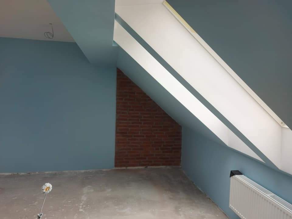 Pokój na poddaszu po świeżym pomalowaniu ścian na niebiesko, z widocznym wałkiem malarskim na podłodze i fragmentem ściany z cegły.