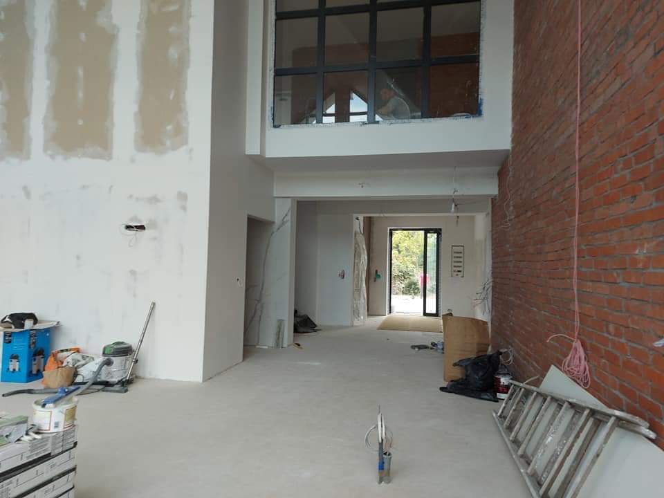 Wnętrze w trakcie remontu: ściana z częściowo położoną płytą kartonowo-gipsową, surowe ściany z cegły, widoczne narzędzia i materiały budowlane, wysokie okno z czarnymi ramami w górnej części...