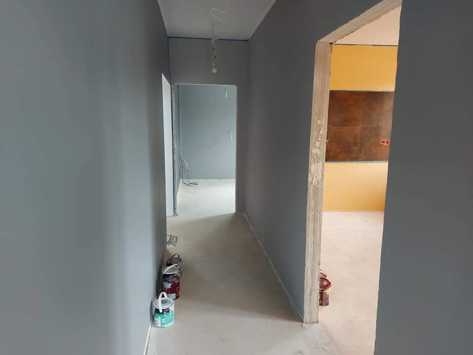 Korytarz w trakcie malowania na szaro, widoczne puszki z farbą przy ścianie, w tle otwarte drzwi do pomalowanego na żółto pokoju z brązowym panelem.
