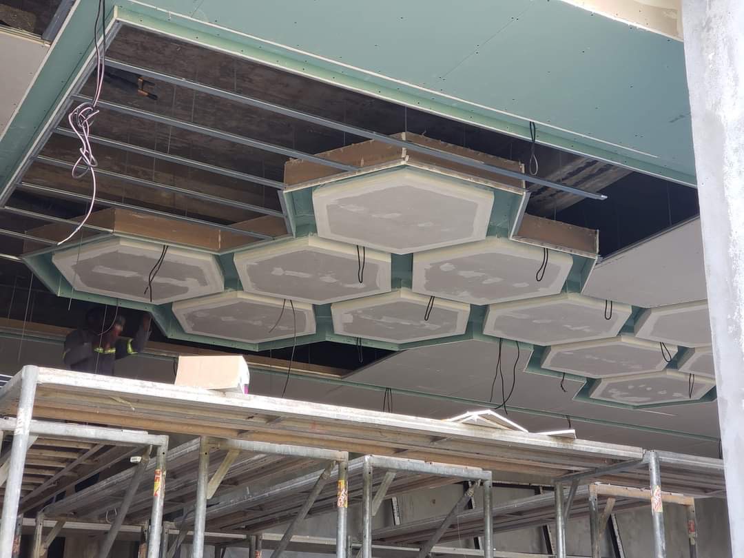 Montaż sufitu podwieszanego z płyt kartonowo-gipsowych w kształcie heksagonów, widoczne metalowe profile konstrukcyjne i instalacja elektryczna, praca w trakcie realizacji.