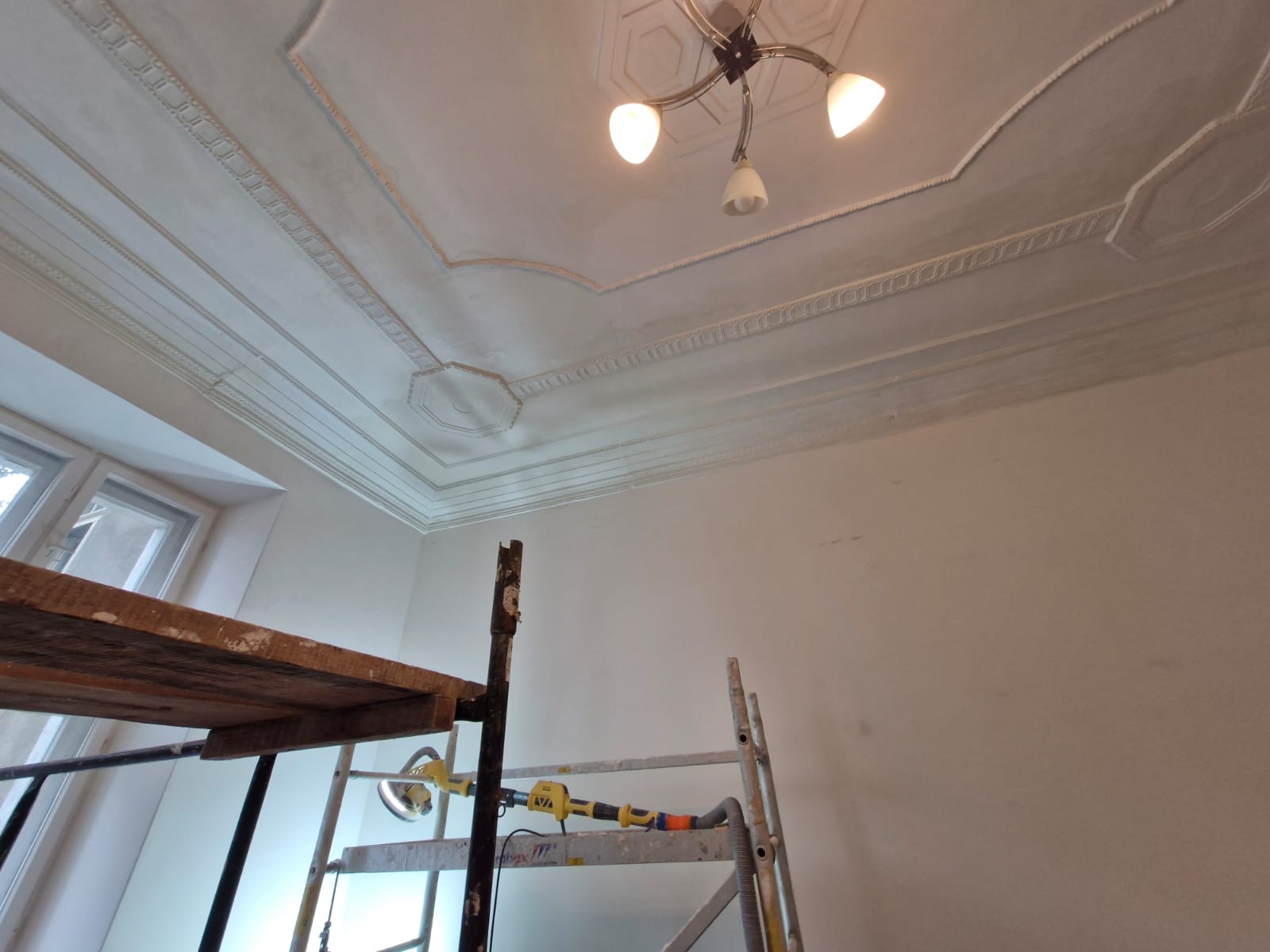 Wnętrze pokoju w trakcie remontu, widoczne rusztowanie, ściana i sufit po szpachlowaniu z dekoracyjnymi sztukateriami, lampa sufitowa z trzema kloszami.