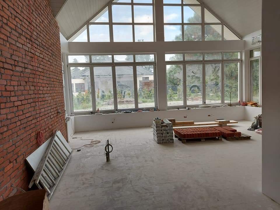 Wnętrze w trakcie prac murarskich: ściana z czerwonej cegły, duże okna panoramiczne z widokiem na ogród, przygotowane cegły na paletach, drabina oparta o ścianę, surowy beton na podłodze.