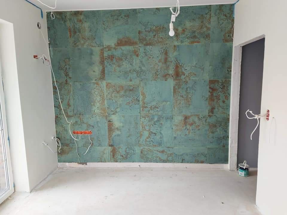 Ściana wyłożona kwadratowymi panelami w odcieniach turkusu i rdzy, surowy beton na podłodze, wystające kable elektryczne i puszki instalacyjne, przygotowania do wykończenia wnętrza.