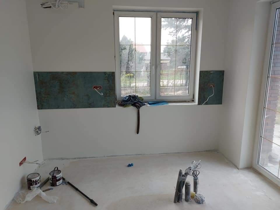 Pokój w trakcie odświeżania: świeżo pomalowane ściany na biało, puszki z farbą, wałek malarski, odsłonięte przewody elektryczne w ścianach, fragment okna z widokiem na zewnątrz.