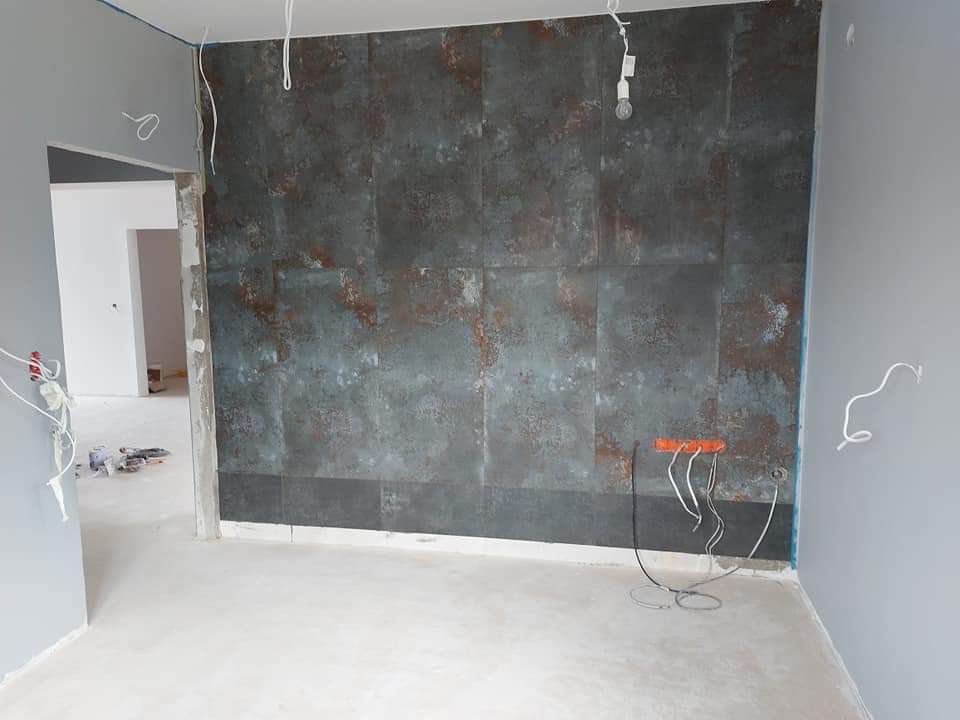 Pokój w trakcie remontu z częściowo wykończoną ścianą z płyt imitujących metal, widoczne kable elektryczne i przygotowane podłoże.