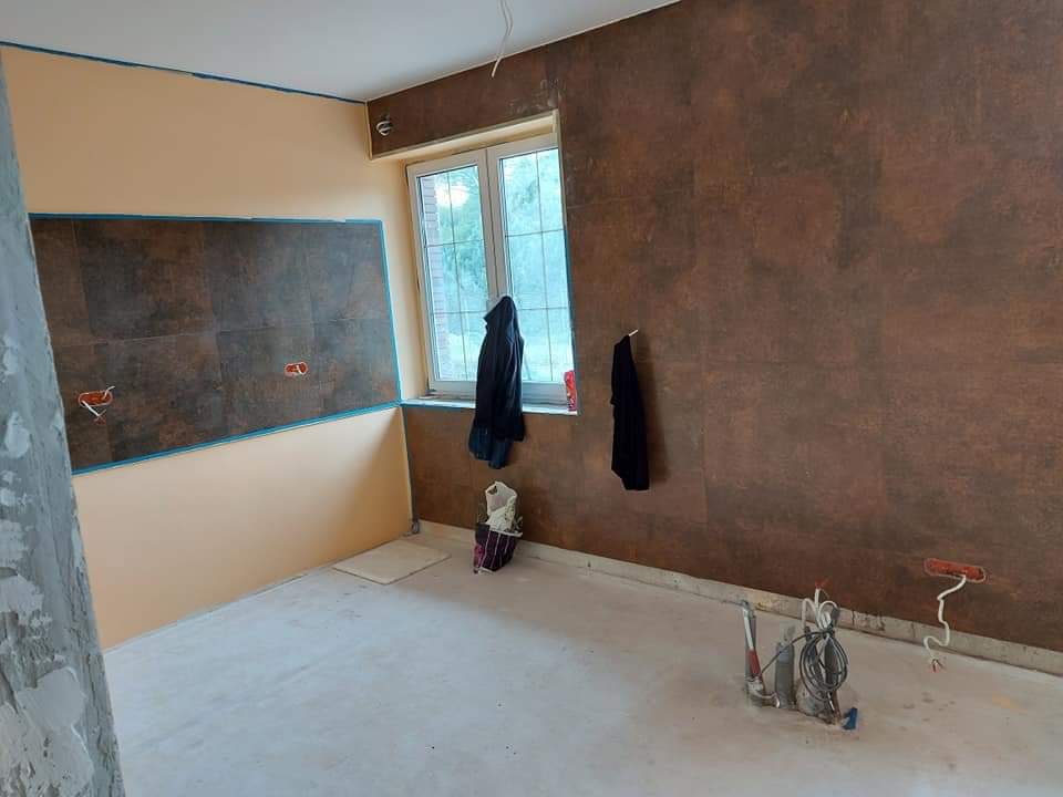 Pokój w trakcie prac wykończeniowych: ściany częściowo pomalowane na beżowo i brązowo z efektem rdzy, widoczne narzędzia malarskie i przewody elektryczne wystające ze ściany, okno z powieszonymi...